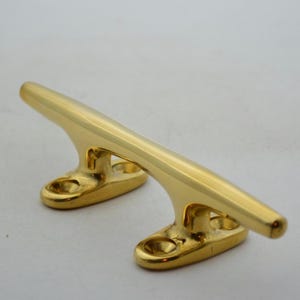 May include: A shiny gold-colored boat cleat. The cleat has a curved top and two mounting bases with holes for screws. The cleat is made of a solid, polished metal and is designed for securing ropes on boats or docks.