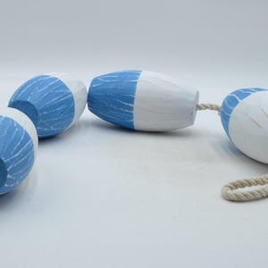 May include: A string of four wooden buoys, painted in blue and white, connected by a natural rope. The buoys have a rounded, oval shape with a textured surface. The blue sections have a carved, linear pattern.