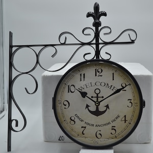 May include: Black metal double-sided wall clock with a white face and black hands. The clock face has the words "Welcome" and "Drop Your Anchor Here" printed on it. The clock is mounted on a black metal bracket with decorative scrolls.