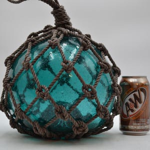 May include: A round, translucent teal glass float encased in a brown knotted rope net. The float is positioned next to a standard 12 fluid ounce aluminum soda can for scale, showing the relative size of the glass sphere and its textured surface.