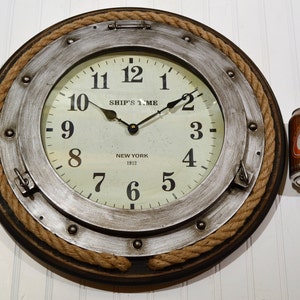 May include: A round wall clock with a nautical theme. The clock face is off-white with black numerals and hands. The clock is surrounded by a silver-colored metal frame, a dark brown wooden frame, and a rope border. The text "SHIP'S TIME NEW YORK 1912" is visible.