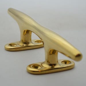 May include: Two polished brass cleats, ideal for marine applications. Each cleat features a curved top and a base with two mounting holes. The cleats are a shiny gold color and are designed for securing ropes or lines.