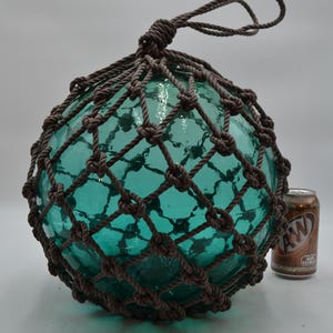 May include: A large, spherical, translucent teal glass fishing float encased in a dark brown knotted rope net. The float features a loop at the top for hanging and is positioned next to a standard 12-ounce A&W Root Beer aluminum soda can.