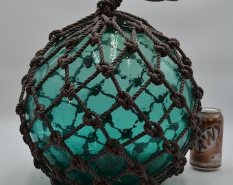 May include: A large, spherical, translucent teal glass fishing float encased in a dark brown knotted rope net. The float features a loop at the top for hanging and is positioned next to a standard 12-ounce A&W Root Beer aluminum soda can.