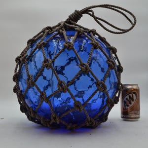 May include: A large, spherical, cobalt blue glass fishing float encased in a brown knotted rope net with a hanging loop at the top. An A&W root beer can is positioned next to the float to provide a visual scale reference for its size.