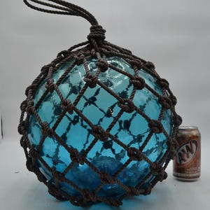 May include: A large, round, blue glass fishing float encased in a brown knotted rope net. The float has a loop at the top for hanging. A brown and tan A&W soda can is next to the float for scale.