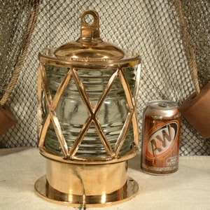May include: A brass nautical light with a clear glass lens and a diamond-shaped wire cage. The light is sitting on a white surface next to a can of A&W Root Beer.