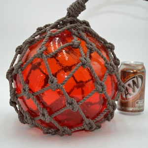 May include: A large, round, orange glass fishing float encased in a brown rope net. The float has a textured surface and is suspended by a rope. An A&W root beer can is next to the float. The float is a decorative nautical item.