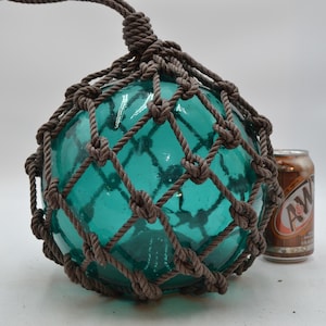 May include: A large, spherical, translucent teal glass fishing float encased in a thick, hand-knotted brown rope net. The float is positioned next to a standard 12-ounce A&W root beer soda can for scale, demonstrating its significant size.