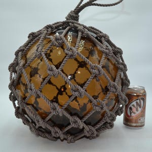 May include: A large, spherical, amber-colored glass float encased in a decorative, hand-knotted gray rope netting. The glass orb features a smooth, reflective surface. An A&W Root Beer soda can is positioned to the right for scale comparison.