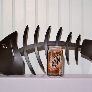 May include: A black metal fish skeleton wall decor with a can of A&W Root Beer in front of it.