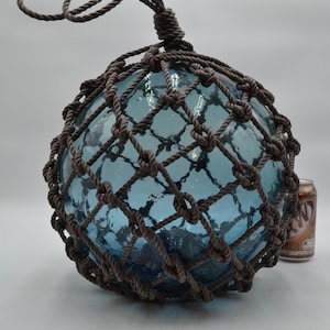 May include: A large, spherical, light blue glass fishing float encased in a dark brown knotted rope net. The glass has a textured, bubbled surface. A standard-sized soda can is positioned to the right for scale, showing the float's size.
