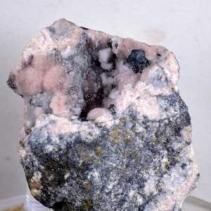 May include: A rock specimen with a vug filled with pink and white crystals. The rock is mostly grey with some gold flecks.