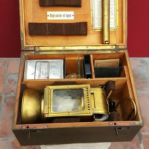 Op de afbeelding: Een open houten kist met een messing lantaarn erin. De kist heeft compartimenten voor verschillende items, waaronder een metalen container, glazen dia's en een klein boekje. De lantaarn heeft een glazen raam en een messing handvat.