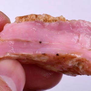 Puede incluir: Primer plano de una piedra preciosa de ópalo rosa. La piedra tiene una apariencia en capas con un tono rosa claro y una superficie rugosa y texturizada. Pequeñas inclusiones oscuras son visibles dentro de la piedra. El borde superior tiene una corteza rugosa de color marrón.