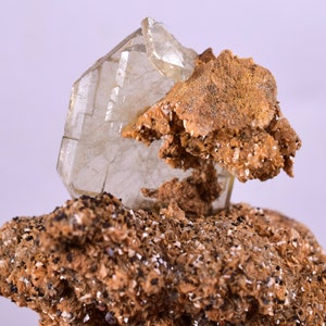 Puede incluir: Un cristal de barita transparente y translúcido con una estructura en capas, anidado en un lecho de roca marrón y beige. El cristal está parcialmente incrustado en la roca, con una parte sobresaliendo.
