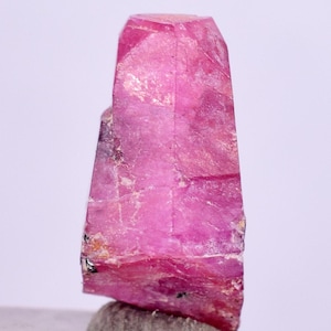 Puede incluir: Un cristal de rubí rosa intenso con una forma puntiaguda facetada. El cristal tiene una textura rugosa y natural y es translúcido.