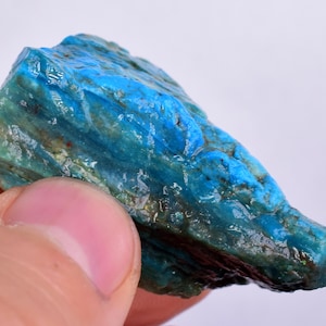 Może przedstawiać: Surowy, niebiesko-zielony kamień szlachetny o warstwowej, prążkowanej strukturze. Kamień ma żywy turkusowy odcień i lekko prześwitujący wygląd.