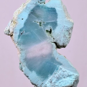 Puede incluir: Una sección transversal de un espécimen mineral, que muestra una mezcla de tonos azul claro y blanco. Los bordes exteriores tienen una apariencia rugosa y texturizada, mientras que el interior revela una estructura más suave y en capas. El espécimen está montado sobre un soporte transparente.