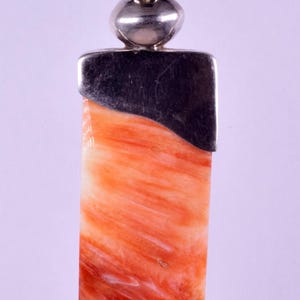 Puede incluir: Un colgante rectangular con una piedra pulida de color naranja y blanco veteado, engastada en una tapa de plata lisa. El colgante está sujeto a una anilla de plata y cuelga de un cordón negro fino, mostrando un degradado de color y textura.