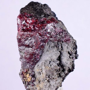 Może przedstawiać: Zbliżenie czerwonego okazu mineralnego z ciemnoszarą macierzą. Minerał ma błyszczący, metaliczny połysk i jest osadzony w macierzy.