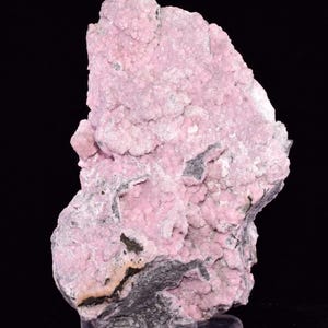 May include: A pink mineral specimen with a rough, textured surface. The rock has a bubbly appearance with areas of dark grey and black. The mineral is displayed on a clear stand against a black background.