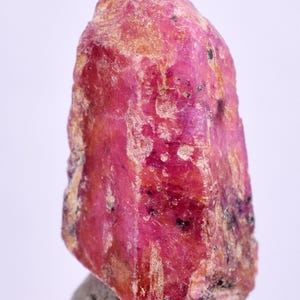 Puede incluir: Un cristal de rubí rojo intenso con una superficie rugosa y natural. El cristal está sobre un pequeño pedestal gris.