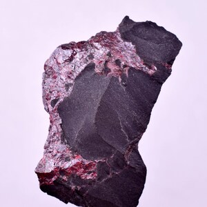 Puede incluir: Una roca gris oscura con una textura rugosa y una sección de depósitos minerales de color púrpura rojizo brillante. La roca tiene una forma irregular y parece ser un espécimen natural, posiblemente un mineral o gema.
