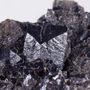 Puede incluir: Primer plano de un grupo de cristales minerales negros. Los cristales tienen una forma geométrica y un brillo metálico. La matriz circundante es un material oscuro y rugoso. La imagen está bien iluminada, resaltando los detalles del espécimen mineral.