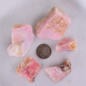 Puede incluir: Cinco piedras de ópalo rosa de diferentes tamaños y formas, con una moneda de diez céntimos de euro para la escala.