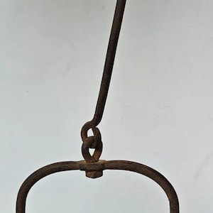 Op de afbeelding: Een antieke, roestige metalen olielamp met een gebogen handvat en een vogelvormig detail aan de bovenkant. De lamp heeft een cirkelvormige basis en een haak om op te hangen. Het metaal heeft een verweerde uitstraling.