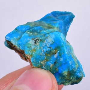 Może przedstawiać: Duży, surowy kawałek turkusu o żywym niebieskim kolorze z zielonymi i białymi wtrąceniami. Turkus jest naturalnym kamieniem i ma unikalny, nieregularny kształt.