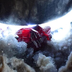 Puede incluir: Primer plano de una vibrante formación cristalina roja, posiblemente un espécimen mineral, sobre un fondo de estructuras cristalinas blancas y grises. El cristal está iluminado, mostrando sus facetas y color.