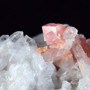 Puede incluir: Primer plano de un espécimen mineral con cristales de cuarzo transparente y una prominente formación de cristal cúbico rosa. Los cristales tienen una textura natural y rugosa, con tonos variables de blanco y rosa.