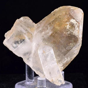 Puede incluir: Un grupo de cristales de cuarzo transparente con un punto central grande y multifacético y cristales más pequeños adjuntos. El cuarzo tiene una apariencia translúcida con algunas áreas de tonos marrón claro y amarillo. El cristal está montado sobre un soporte transparente sobre un fondo negro.