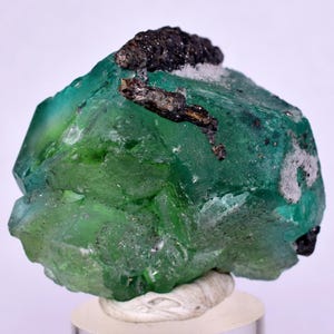 May include: A vivid green mineral specimen with dark, irregular inclusions and a white base. The gem-quality stone has a crystalline structure and is displayed on a clear stand.