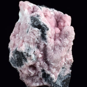 May include: A close-up of a mineral specimen with light pink, crystalline formations. The pink crystals are contrasted by dark grey and black rock. The specimen is set against a black background.