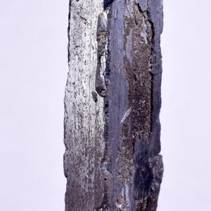 Puede incluir: Un gran espécimen mineral metálico gris oscuro con una superficie rugosa y texturizada. El mineral tiene una forma rectangular alargada y parece ser un solo cristal.