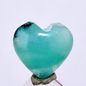 Puede incluir: Una gema pulida en forma de corazón, de color turquesa vibrante. La piedra tiene una superficie lisa y brillante con una pequeña muesca en la parte superior. Está montada sobre una pequeña base de textura rugosa, sobre un fondo blanco.