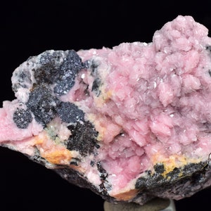 May include: A pink and grey mineral specimen with a rough, textured surface. The pink crystals are clustered together and have a shiny, translucent appearance. The grey areas are made of a different mineral and have a more matte finish.