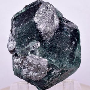 May include: A dark green mineral specimen with patches of silver-coloured metallic material. The crystalline structure is visible, with sharp edges and a rough texture. The mineral sits on a clear acrylic stand.