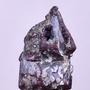 May include: A close-up of a mineral specimen with a dark red and silver crystalline structure. The mineral has a rough, textured surface with areas of deep red and metallic silver. The base is a light grey colour.