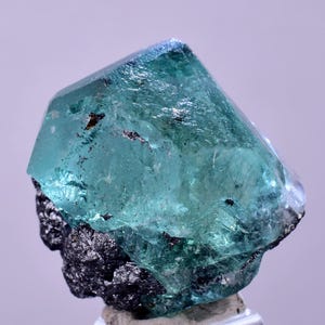 May include: A teal crystal with a geometric shape, set on a dark, rough mineral base. The crystal's surface has a textured appearance, with areas of transparency. The crystal's colour is a vibrant teal.