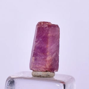 Puede incluir: Un cristal rosa facetado y alargado con una superficie rugosa y texturizada. El cristal está montado sobre una pequeña base redonda gris. El color del cristal varía de rosa claro a oscuro.