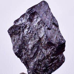 Puede incluir: Un espécimen mineral de color gris oscuro, metálico, con textura rugosa. El mineral tiene un ligero brillo iridiscente.