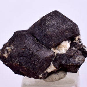 Puede incluir: Un grupo de cristales de color gris oscuro, casi negro, con una textura ligeramente rugosa. Los cristales están entrelazados y tienen una forma cúbica. Se puede ver algo de material cristalino blanco en el centro del grupo.