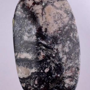 May include: An oval-shaped polished stone cabochon featuring a complex marbled pattern of deep black, charcoal grey, and creamy white veins. The surface is smooth and glossy, displaying a natural mineral texture with varied dark and light inclusions.