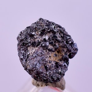 Może przedstawiać: Ciemny, nieregularny kształt okazu mineralnego o szorstkiej, teksturowanej powierzchni. Minerał jest głównie ciemnoszary i czarny, z niektórymi obszarami o metalicznym połysku. Podstawa ma jaśniejszy, brązowy kolor.