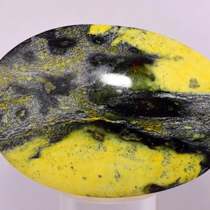 Pode incluir: Gema oval com um padrão marcante de amarelo, preto e cinza. A pedra polida exibe uma mistura de cores e texturas, com uma superfície brilhante. A gema é provavelmente usada para joias ou fins decorativos.
