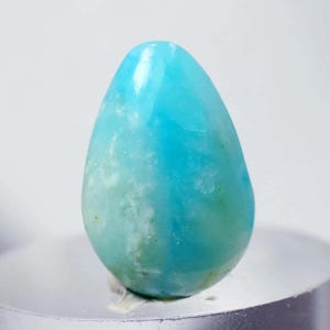 Puede incluir: Una gema pulida en forma de lágrima, en tonos turquesa y azul aguamarina. La piedra tiene una superficie lisa y brillante con sutiles inclusiones blancas y verdes. La gema se muestra sobre una superficie plateada sobre un fondo blanco.